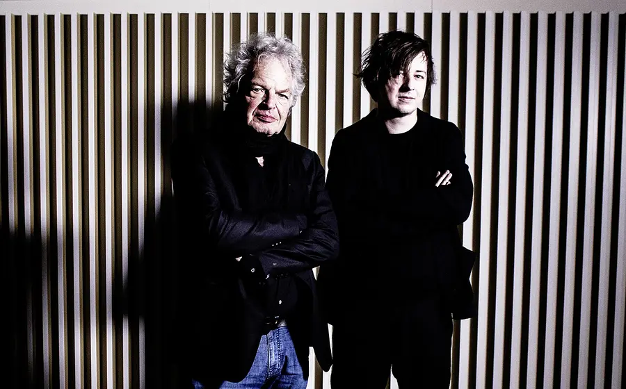 Michael Wollny und Joachim Kühn vor einer Wand mit vertikalen Lamellen, beide in dunkler Kleidung mit verschränkten Armen vor kontrastreicher Beleuchtung