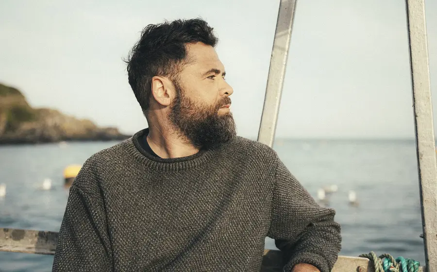 Der Künstler Passenger mit dunklem Haar und Vollbart in grauem Strickpullover auf einem Boot, mit Blick zur Seite vor Küstenlandschaft und ruhigem Meer im Hintergrund.
