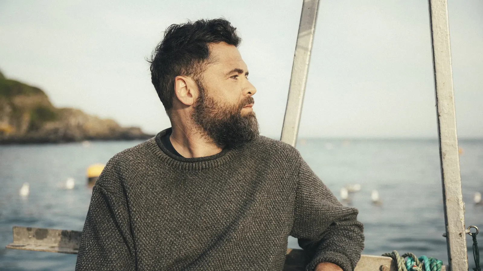 Der Künstler Passenger mit dunklem Haar und Vollbart in grauem Strickpullover auf einem Boot, mit Blick zur Seite vor Küstenlandschaft und ruhigem Meer im Hintergrund.