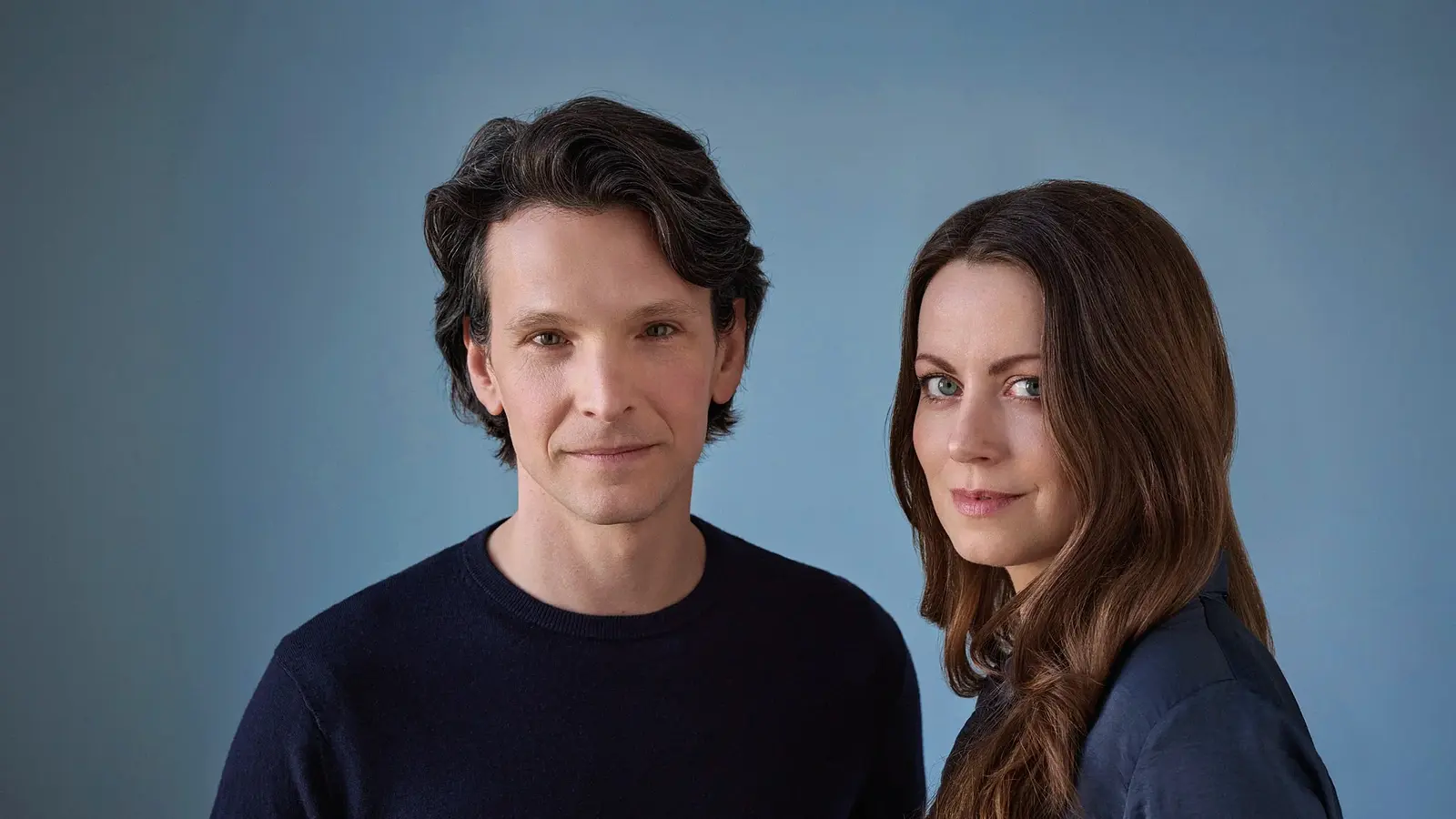 Sabin Tabrea und Alice Dwyer lächelnd in dunkler Kleidung vor blauem Hintergrund