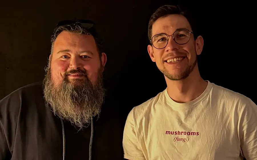 Zwei Personen vor dunklem Hintergrund; die eine trägt ein dunkles Oberteil mit langem Bart, die andere eine Brille und ein T‑Shirt mit der Aufschrift mushrooms - fungi, warm beleuchtet.