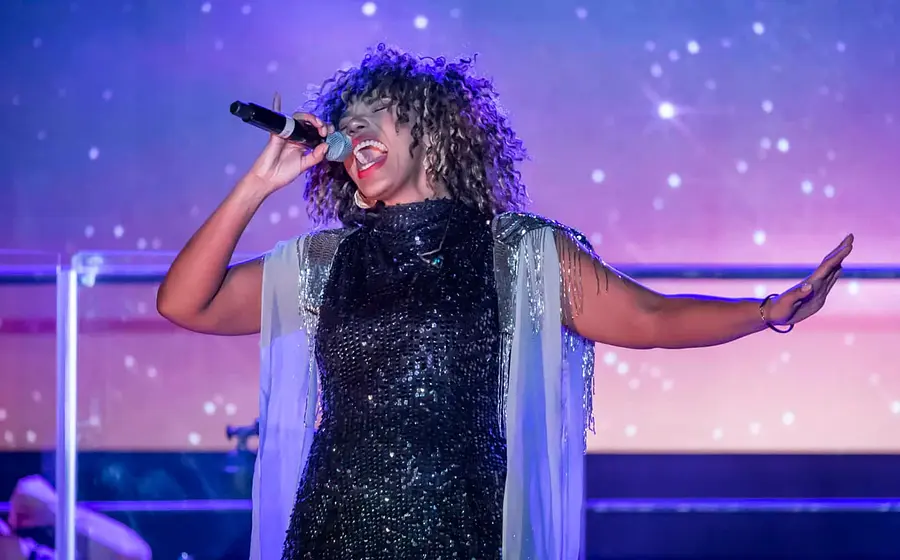 Darstellerin von TINA – The Rock Legend singt mit Mikrofon auf einer Konzertbühne vor farbig beleuchtetem Hintergrund.