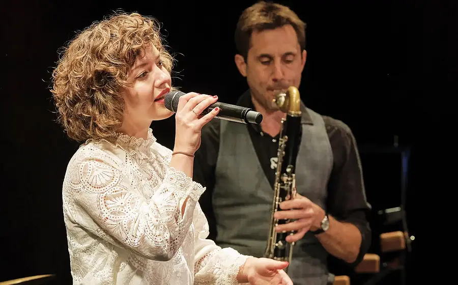 Bühnenfoto Sarah und das Nix mit Person im weißen Spitzenoberteil, die in ein Mikrofon singt, während im Hintergrund eine weitere Person Klarinette spielt, vor dunklem Hintergrund