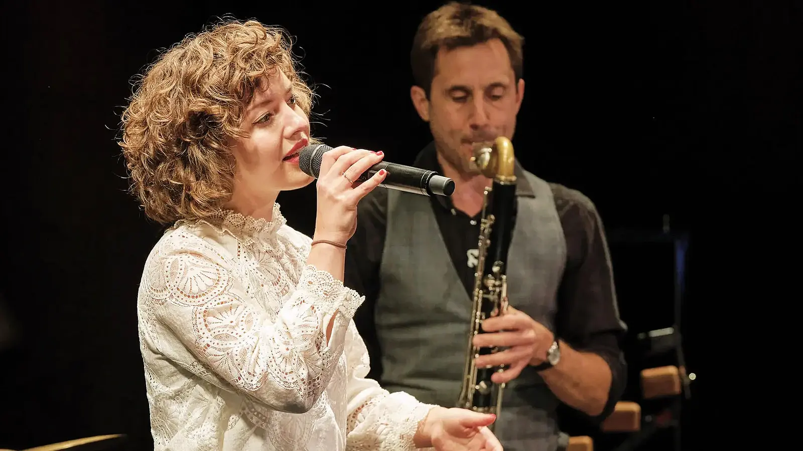 Bühnenfoto Sarah und das Nix mit Person im weißen Spitzenoberteil, die in ein Mikrofon singt, während im Hintergrund eine weitere Person Klarinette spielt, vor dunklem Hintergrund