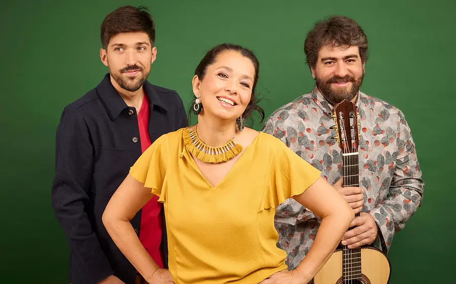 Studiofoto mit drei Personen der Gruppe Amaré vor grünem Hintergrund, eine Person vorne in gelbem Oberteil, daneben zwei weitere Personen, eine mit Gitarre in der Hand