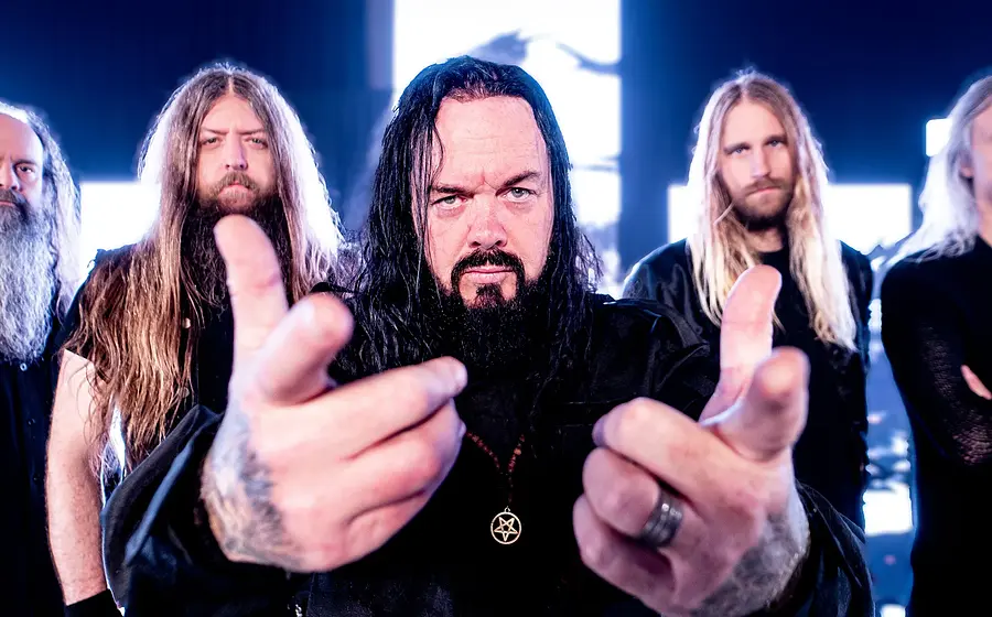 Bandfoto von Evergrey mit fünf Bandmitgliedern in schwarzer Kleidung vor hell leuchtenden Bildschirmen und Instrumenten auf einer Bühnenkulisse, eine Person im Vordergrund mit ausgestreckten Händen und Pentagramm-Anhänger, die weiteren Mitglieder dahinter in einer Reihe angeordnet.