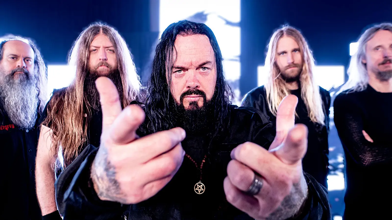 Bandfoto von Evergrey mit fünf Bandmitgliedern in schwarzer Kleidung vor hell leuchtenden Bildschirmen und Instrumenten auf einer Bühnenkulisse, eine Person im Vordergrund mit ausgestreckten Händen und Pentagramm-Anhänger, die weiteren Mitglieder dahinter in einer Reihe angeordnet.