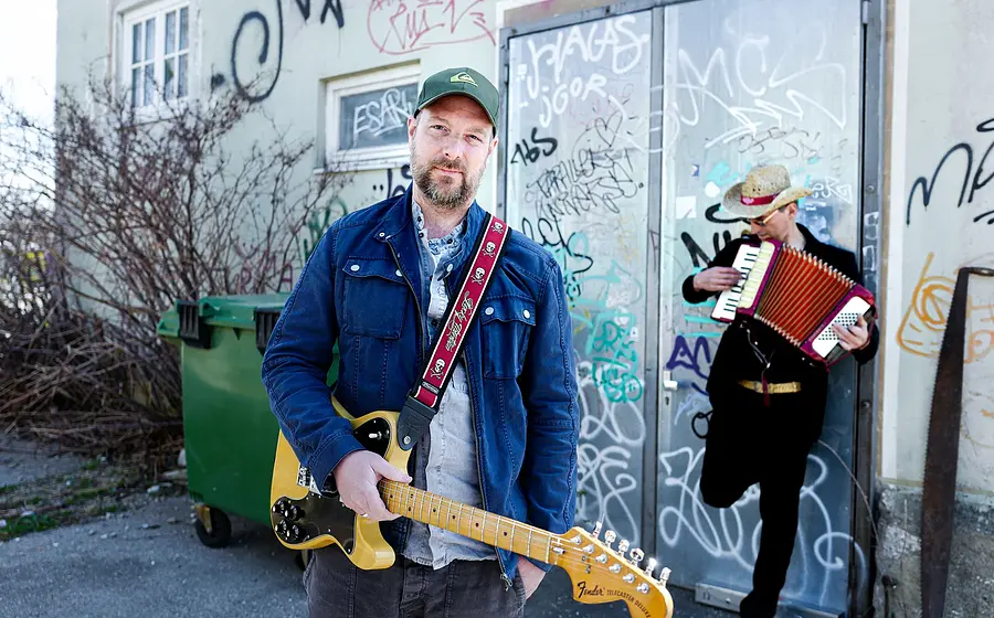 Der Künstler mit Gitarre vor einer Hauswand mit Graffiti, an der ein weiterer Musiker lehnt