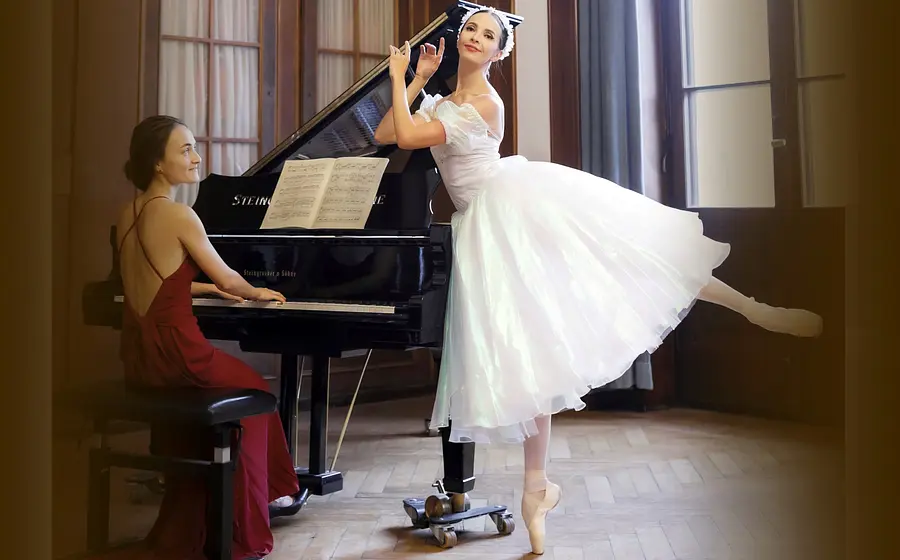 Eine Pianistin im roten Abendkleid am Flügel und eine Balletttänzerin im weißen Tüllkleid auf Spitze, in einem historischen Saal mit hohen Fenstern und geöffnetem Konzertflügel.
