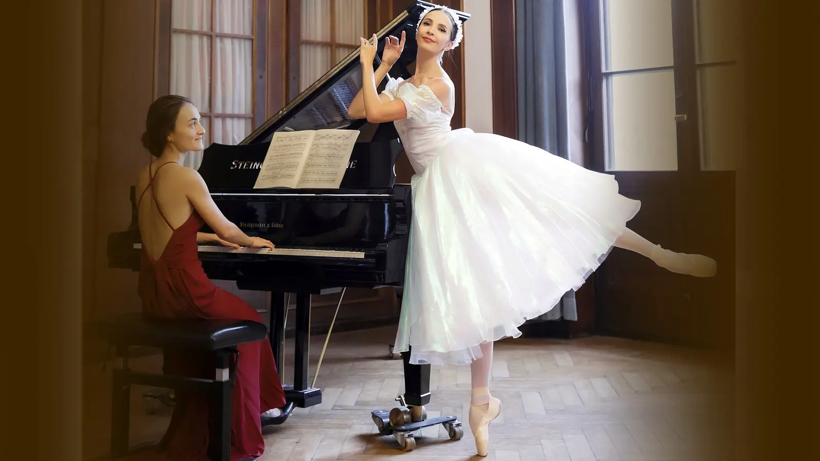 Eine Pianistin im roten Abendkleid am Flügel und eine Balletttänzerin im weißen Tüllkleid auf Spitze, in einem historischen Saal mit hohen Fenstern und geöffnetem Konzertflügel.