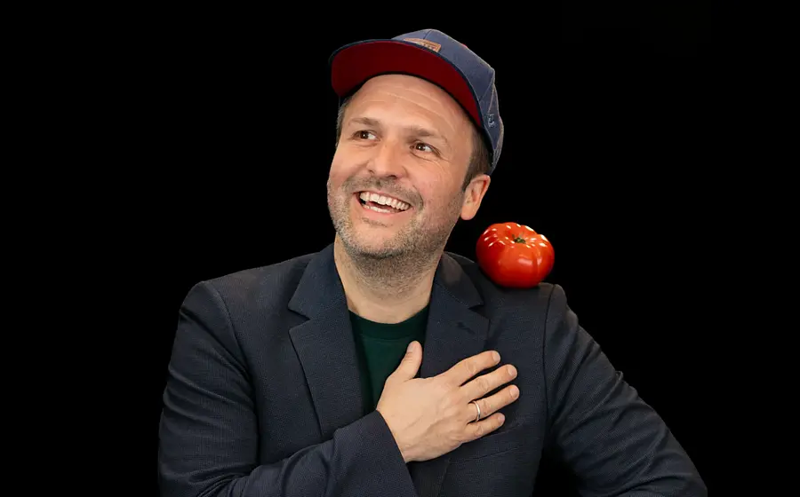Alt-Text (kurz und präzise): Porträt eines Mannes mit Kappe und Sakko vor schwarzem Hintergrund, auf der Schulter balanciert eine rote Tomate