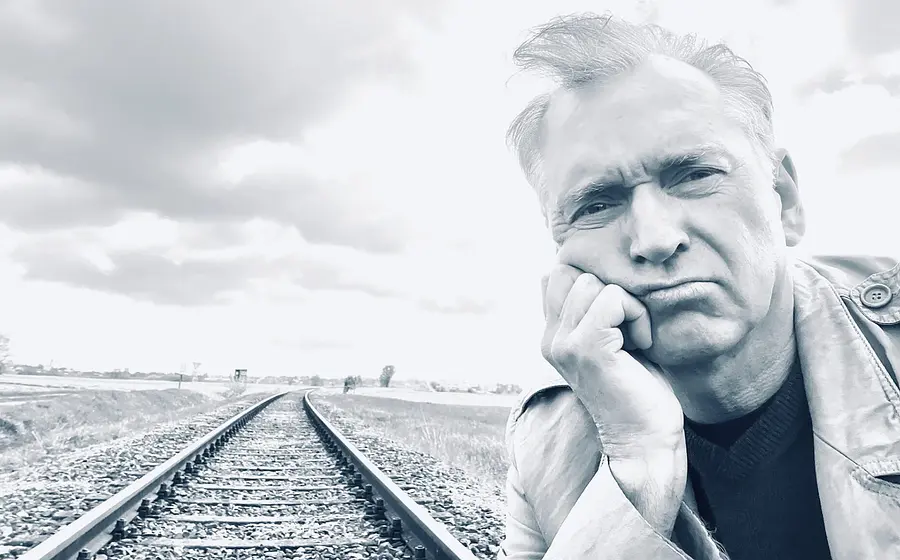 Porträtfoto für eine Veranstaltungsankündigung: Eine Person steht im Vordergrund eines Schwarz‑Weiß‑Motivs und lehnt mit dem Kopf auf der Hand an. Im Hintergrund verlaufen Eisenbahnschienen in die Ferne durch eine offene Landschaft unter bewölktem Himmel. Die Bildkomposition wirkt ruhig und reduziert und eignet sich als Motiv für eine Bühnen‑ oder Kabarettveranstaltung.