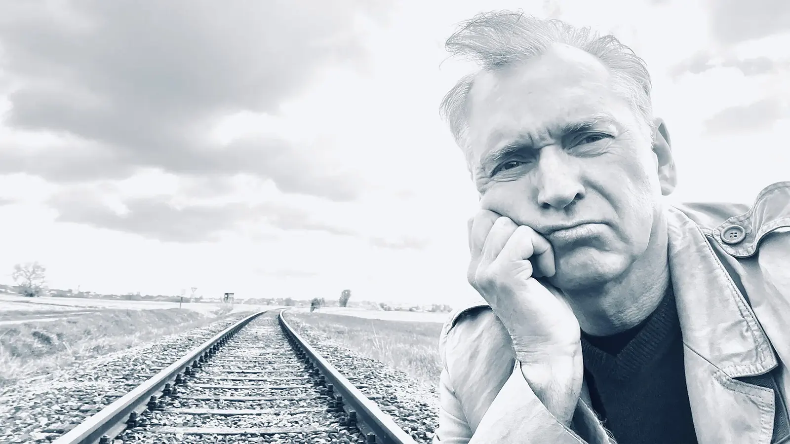 Porträtfoto für eine Veranstaltungsankündigung: Eine Person steht im Vordergrund eines Schwarz‑Weiß‑Motivs und lehnt mit dem Kopf auf der Hand an. Im Hintergrund verlaufen Eisenbahnschienen in die Ferne durch eine offene Landschaft unter bewölktem Himmel. Die Bildkomposition wirkt ruhig und reduziert und eignet sich als Motiv für eine Bühnen‑ oder Kabarettveranstaltung.