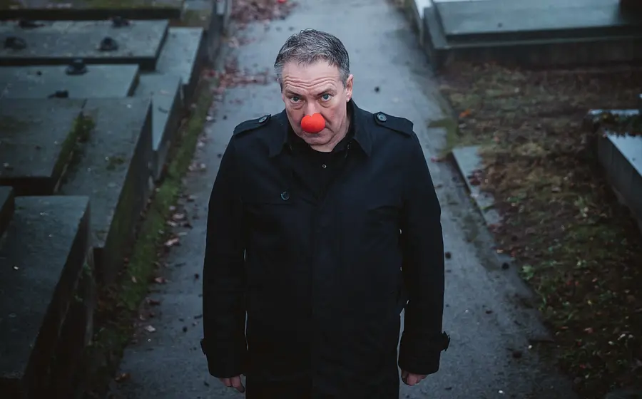 Robert Palfrader mit einer roten Clownsnase in einem schwarzen Mantel auf einem dunklen Weg, links und rechts schwarze  Steinplatten
