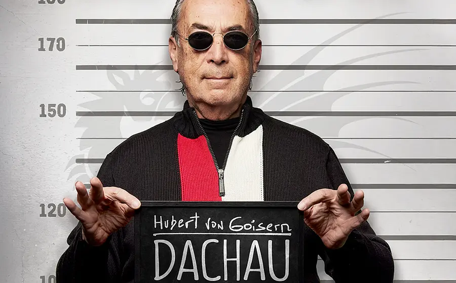 Mann mit Sonnenbrille hält eine Tafel mit der Aufschrift Hubert von Goisern – Dachau vor einer Höhenmarkierung, inszeniert wie ein Polizeifoto