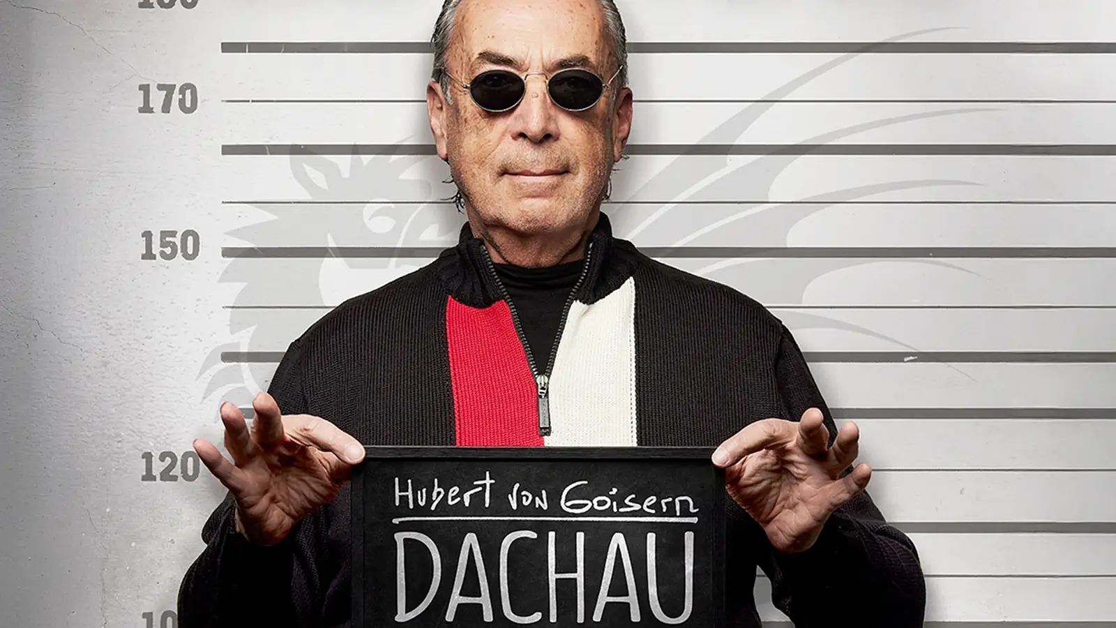 Mann mit Sonnenbrille hält eine Tafel mit der Aufschrift Hubert von Goisern – Dachau vor einer Höhenmarkierung, inszeniert wie ein Polizeifoto