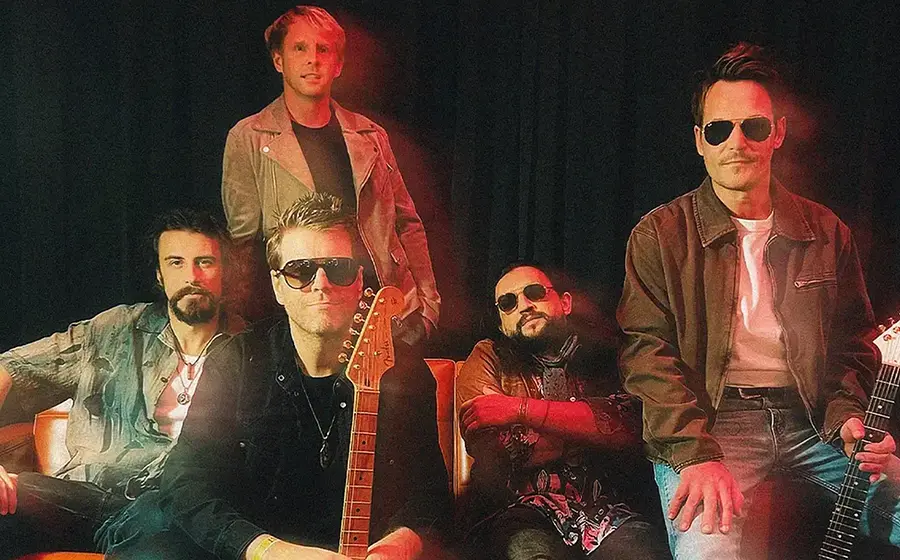 Der Künstler Danny Bryant mit Band auf einem Sofa, einige Personen tragen Sonnenbrillen, in farbigem Scheinwerferlicht vor einem dunklen Vorhang