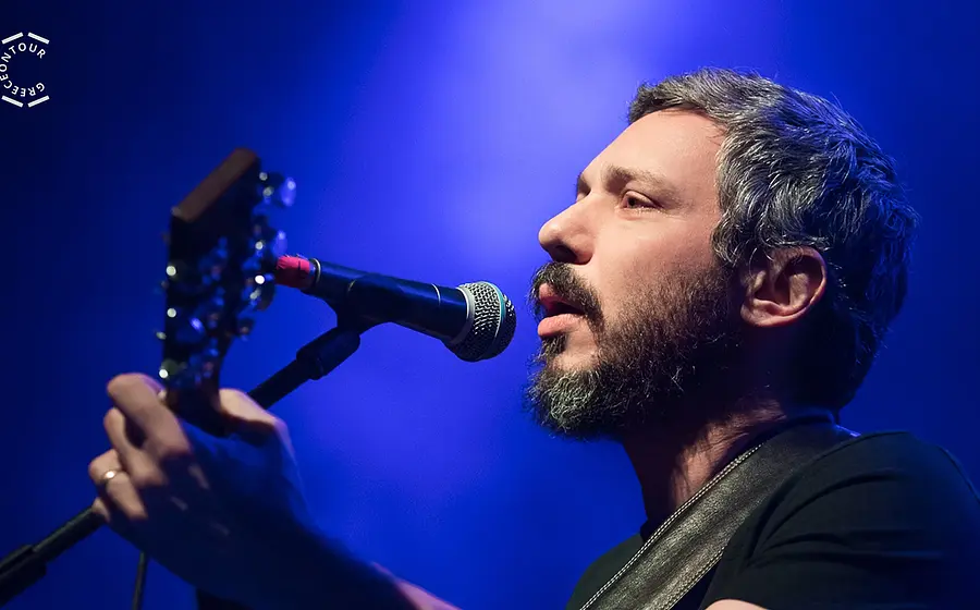 Person mit Gitarre singt auf einer Bühne in ein Mikrofon. Blaue Konzertbeleuchtung und Nahaufnahme während eines Live-Auftritts.