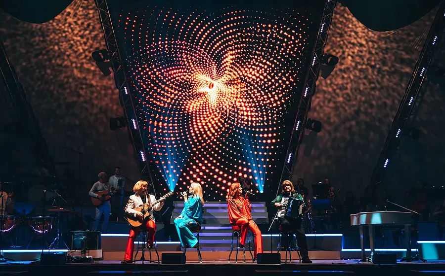Mehrere Künstler der Show **ABBAMANIA THE SHOW** sitzen und performen auf einer beleuchteten Bühne, begleitet von Live-Musikern; im Hintergrund eine große LED-Lichtinstallation mit rot leuchtendem, spiralförmigem Muster.