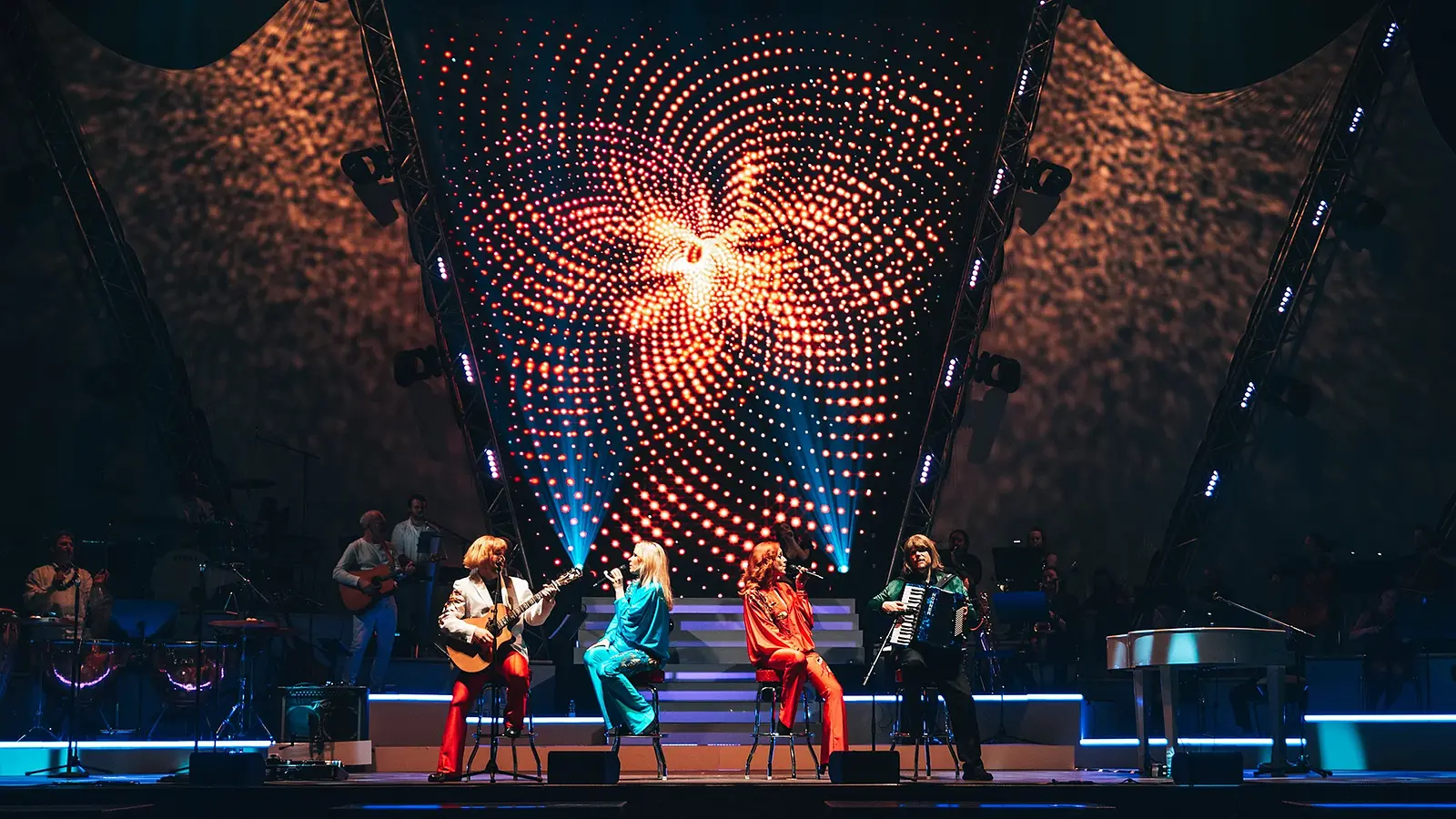 Mehrere Künstler der Show **ABBAMANIA THE SHOW** sitzen und performen auf einer beleuchteten Bühne, begleitet von Live-Musikern; im Hintergrund eine große LED-Lichtinstallation mit rot leuchtendem, spiralförmigem Muster.