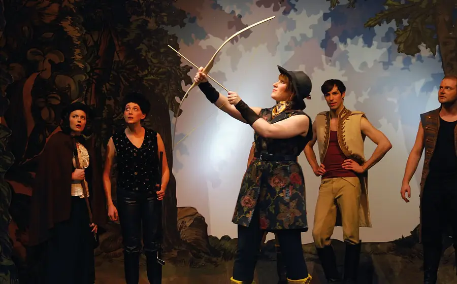 Mehrere Darsteller in Märchenkostümen stehen auf einer Theaterbühne in Waldkulisse. Im Mittelpunkt hält eine Person einen Bogen in die Höhe.
