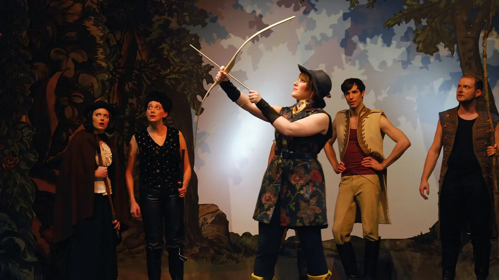 Mehrere Darsteller in Märchenkostümen stehen auf einer Theaterbühne in Waldkulisse. Im Mittelpunkt hält eine Person einen Bogen in die Höhe.