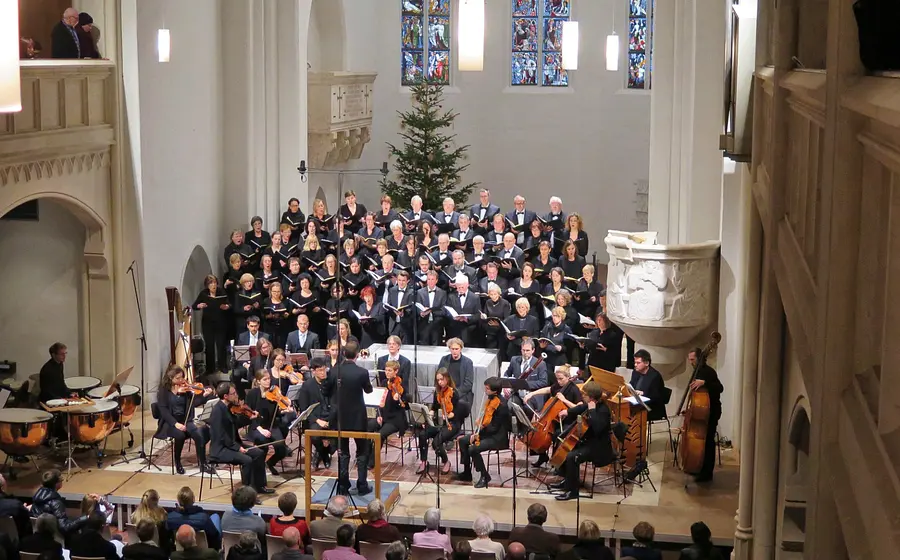 Konzertaufführung eines Chors mit Orchester in einer Kirche. Vor einem großen Chor auf mehreren Podesten dirigiert eine Person das Orchester mit Streichinstrumenten, Pauken und Kontrabass. Im Hintergrund sind ein Weihnachtsbaum sowie farbige Kirchenfenster zu sehen.