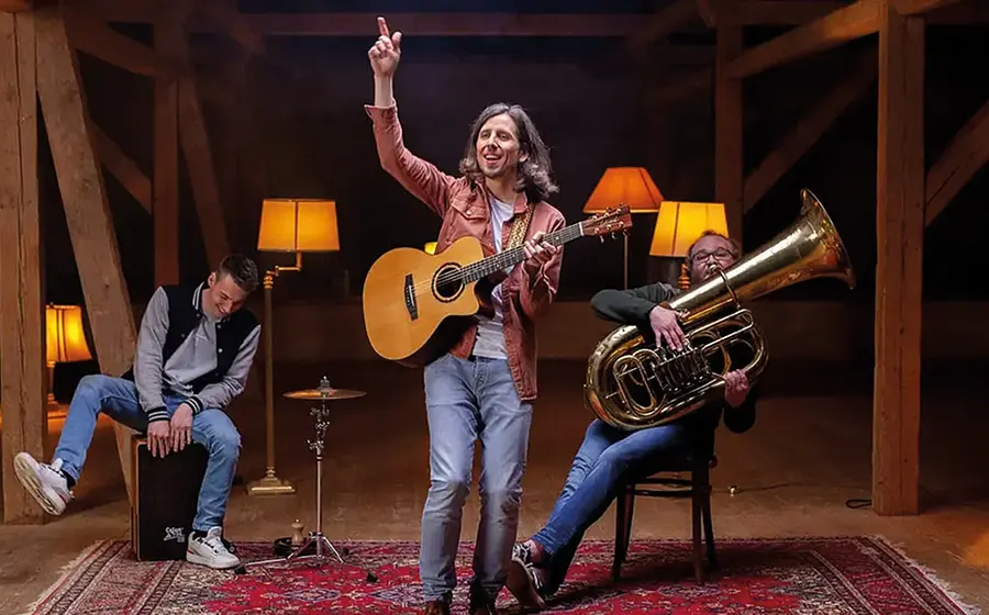 Bandfoto von Jannemacher mit drei Musikern in einer Scheune oder Holzhalle auf orientalischem Teppich, eine Person mit Akustikgitarre im Zentrum, links eine Person auf einer Cajón und rechts eine Person mit Tuba, umgeben von warm beleuchteten Stehlampen und Holzbalken.