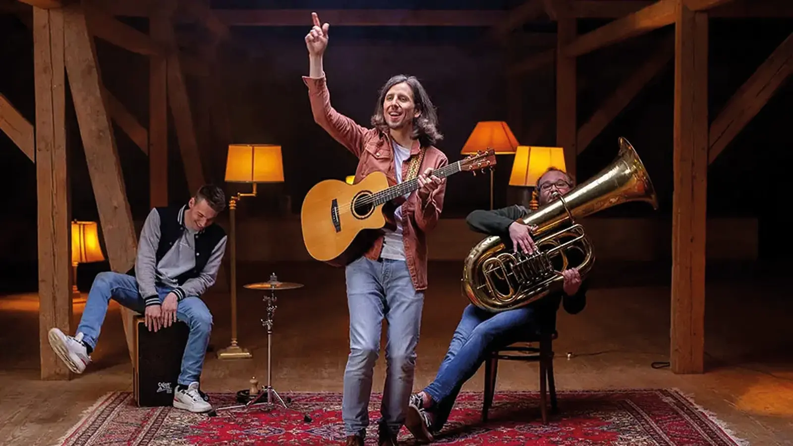Bandfoto von Jannemacher mit drei Musikern in einer Scheune oder Holzhalle auf orientalischem Teppich, eine Person mit Akustikgitarre im Zentrum, links eine Person auf einer Cajón und rechts eine Person mit Tuba, umgeben von warm beleuchteten Stehlampen und Holzbalken.