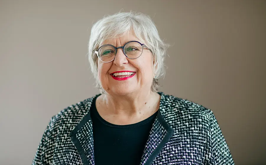 Eine lachende Dame mit weißem Haar, Brille und schwarz-weisser Jacke vor hellbraunem Hintergrund zur Veranstaltung Ladies Lunch