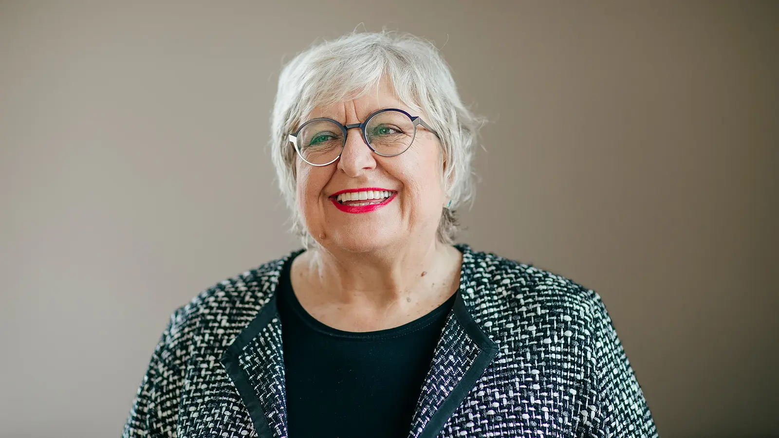 Eine lachende Dame mit weißem Haar, Brille und schwarz-weisser Jacke vor hellbraunem Hintergrund zur Veranstaltung Ladies Lunch