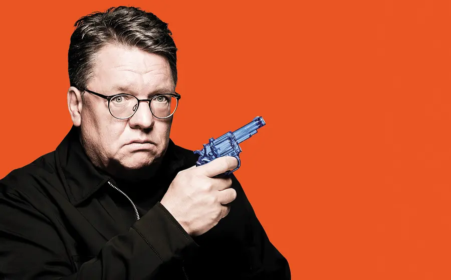 Porträt von Helmut Schleich mit Brille vor orangefarbenem Hintergrund. Die Person hält einen blauen Spielzeugrevolver in der Hand und blickt direkt in die Kamera.