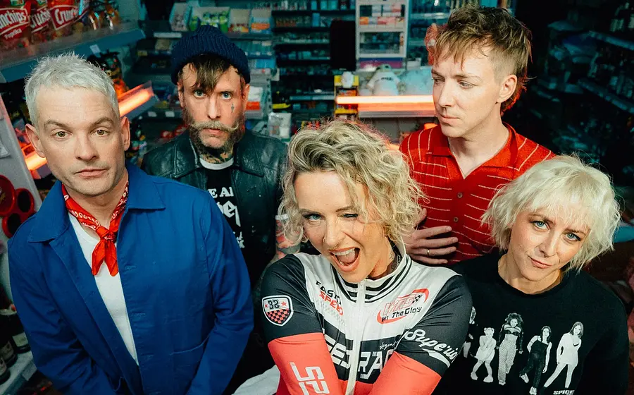 Bandfoto von Jennifer Rostock: Fünf Personen stehen in einem Kiosk zwischen Regalen und blicken in die Kamera. Die Szene ist farbenfroh ausgeleuchtet und wirkt modern und dynamisch.