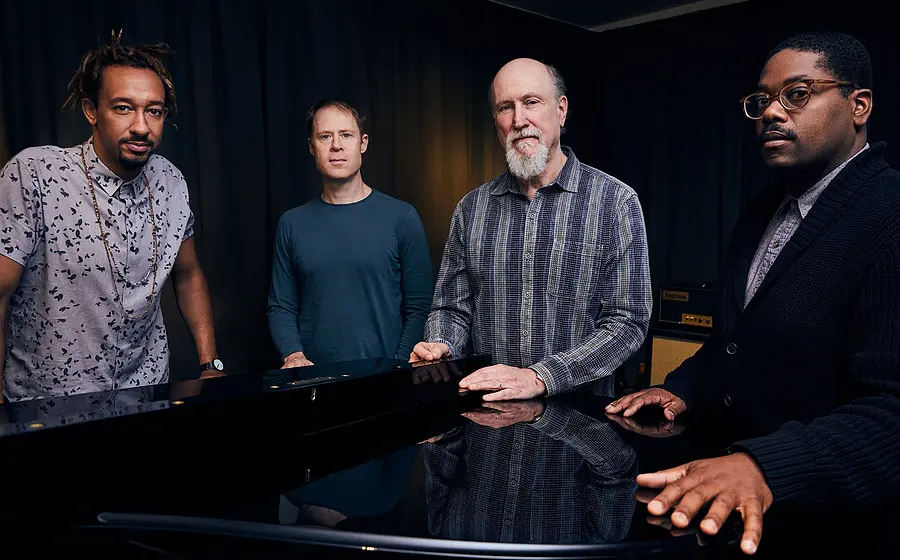 Foto für das Event John Scofield Combo 75 mit der Gruppe, die sich an ein Klavier anlehnt.
