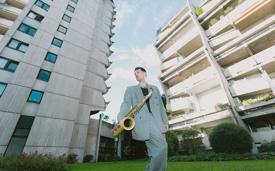 Der Musiker Moritz Stahl mit Saxophon auf einer Wiese zwischen zwei Hochhäusern