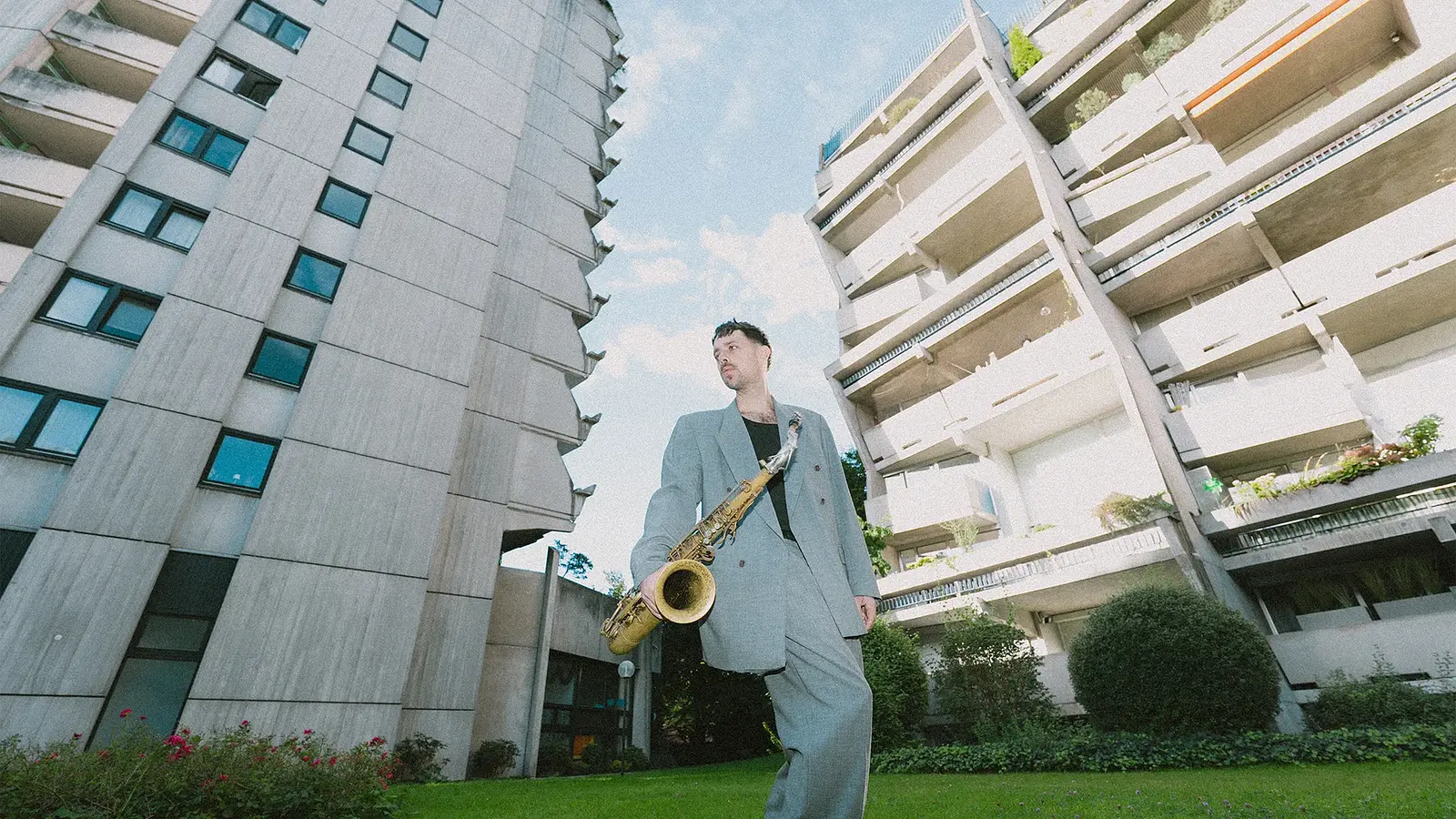 Der Musiker Moritz Stahl mit Saxophon auf einer Wiese zwischen zwei Hochhäusern