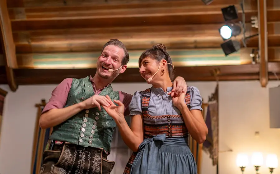 Bühnenfoto vom Kathreintanz mit zwei Personen in bayerischer Tracht, eine in grün gemusterter Weste und Lederhose, die andere im blauen Dirndl, auf einer Bühne mit Headsetmikrofonen und warm beleuchtetem Saal im Hintergrund