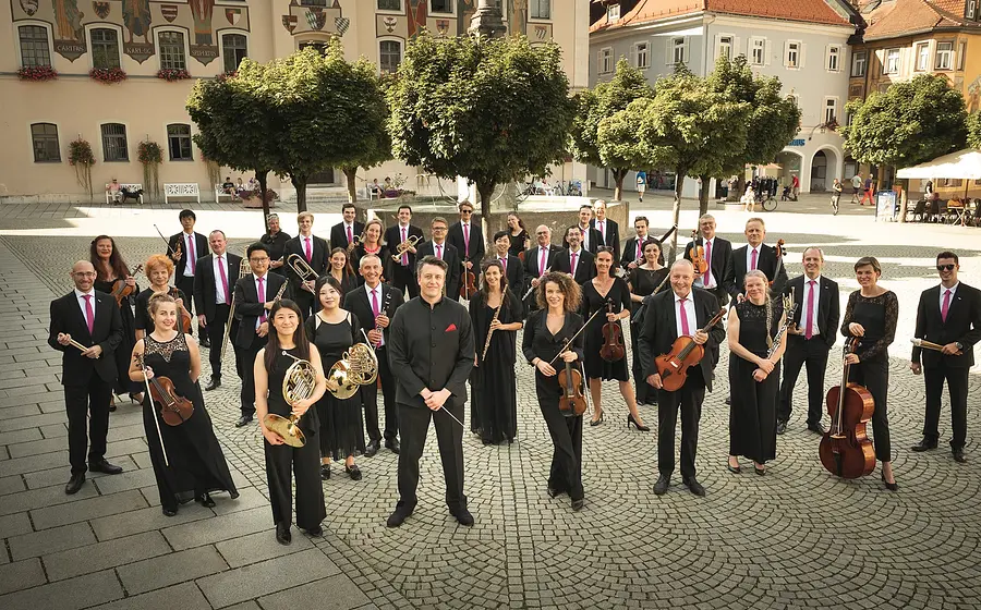 Gruppenfoto eines Orchesters auf einem historischen Marktplatz. Die Musiker tragen schwarze Konzertkleidung mit pinkfarbenen Krawatten oder Tüchern und halten ihre Instrumente vor hellen Gebäuden, grünen Bäumen und warmem Sonnenlicht.