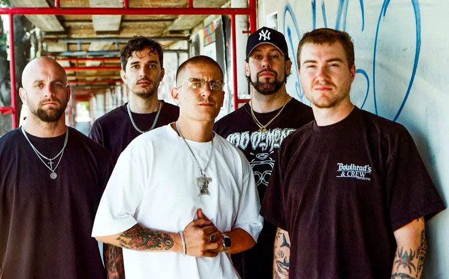 Bandfoto von Pain Of Truth in dunkler Freizeitkleidung vor einer betonierten Passage mit roten Gerüststangen und Graffiti, eine Person im weißen T-Shirt im Vordergrund sowie eine Person mit Baseballkappe im Hintergrund.