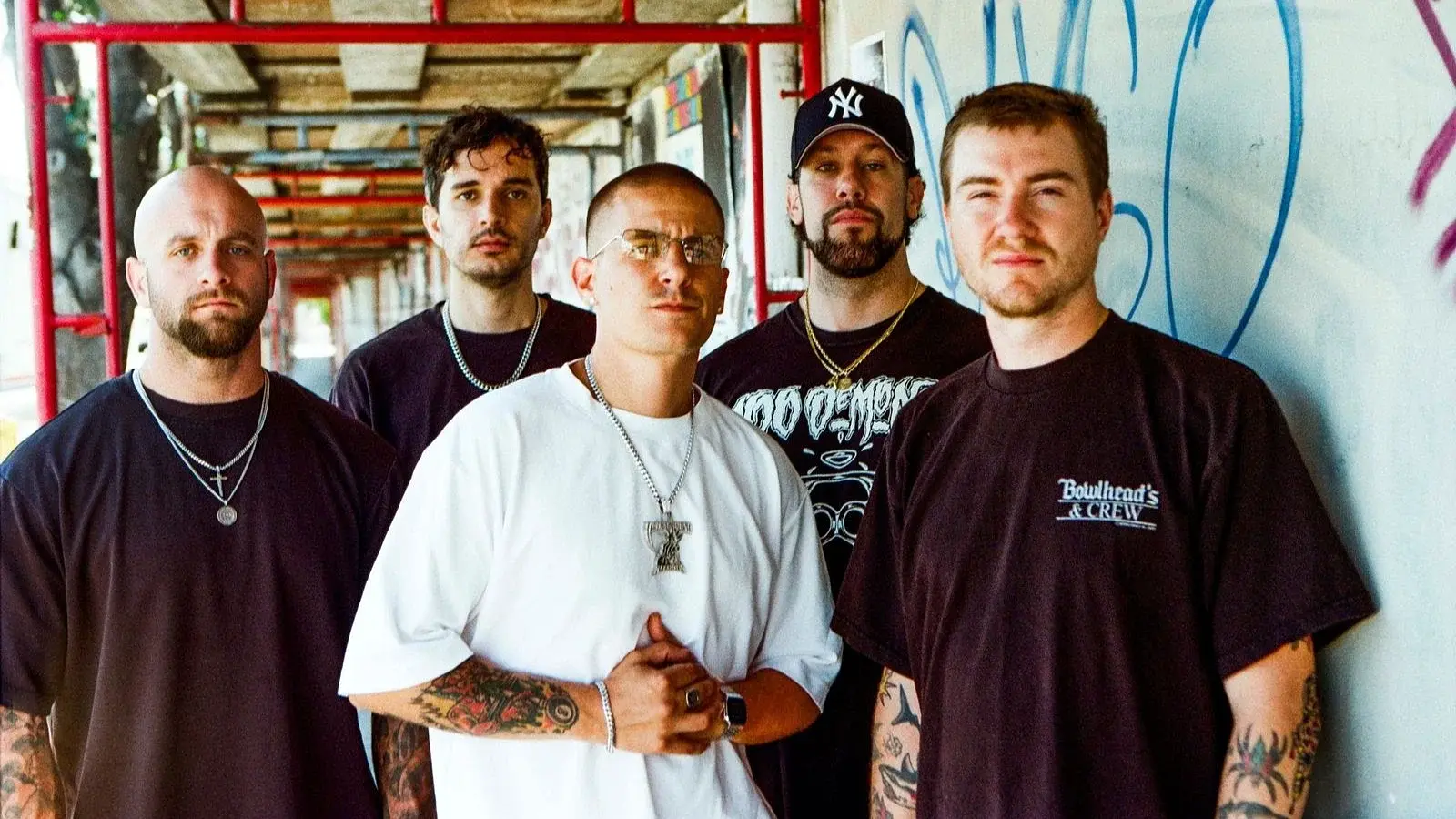 Bandfoto von Pain Of Truth in dunkler Freizeitkleidung vor einer betonierten Passage mit roten Gerüststangen und Graffiti, eine Person im weißen T-Shirt im Vordergrund sowie eine Person mit Baseballkappe im Hintergrund.