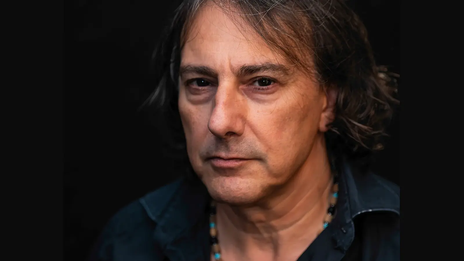 Ein Portrait des Künstlers Pippo Polina in Jeanshemd mit Kette vor dunklem Hintergrund