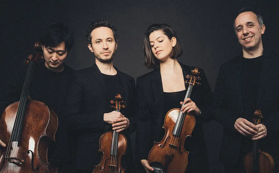 Quatuor Ébène mit vier Musiker:innen in schwarzer Kleidung vor dunklem Hintergrund, jeweils mit Streichinstrumenten in stehender Haltung, die Instrumente werden vor dem Körper gehalten.