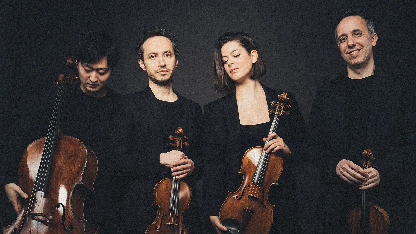Quatuor Ébène mit vier Musiker:innen in schwarzer Kleidung vor dunklem Hintergrund, jeweils mit Streichinstrumenten in stehender Haltung, die Instrumente werden vor dem Körper gehalten.