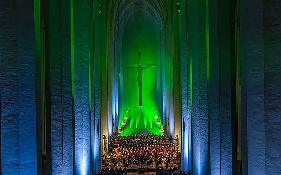 EIn Chor mit Publikum vor dem grün und blau beleuchteten Altar im Innenraum einer Kirche,im Hintergrund ein Jesuskreuz