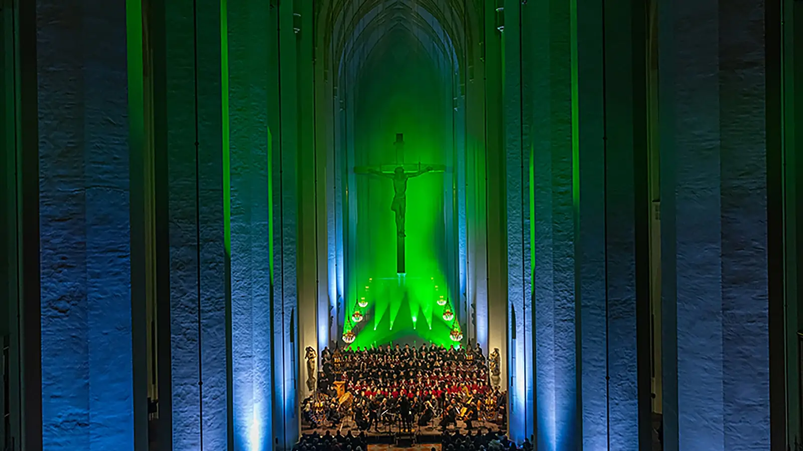 EIn Chor mit Publikum vor dem grün und blau beleuchteten Altar im Innenraum einer Kirche,im Hintergrund ein Jesuskreuz