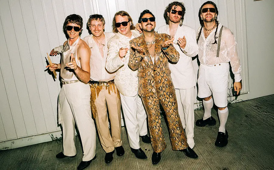 Die sechs Mitglieder von Roy Bianco und die Abbrunzati Boys stehen vor einen weißen Mauer. Sie tragen weiß-beige Bühnenoutfits und Sonnenbrillen.