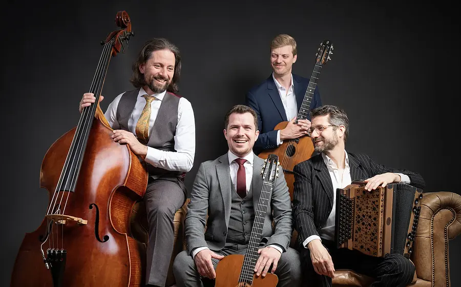 Vier Herren in klassischen Anzügen sitzen mit Instrumenten lachend auf einem braunen Ledersofa vor dunklem Hintergrund
