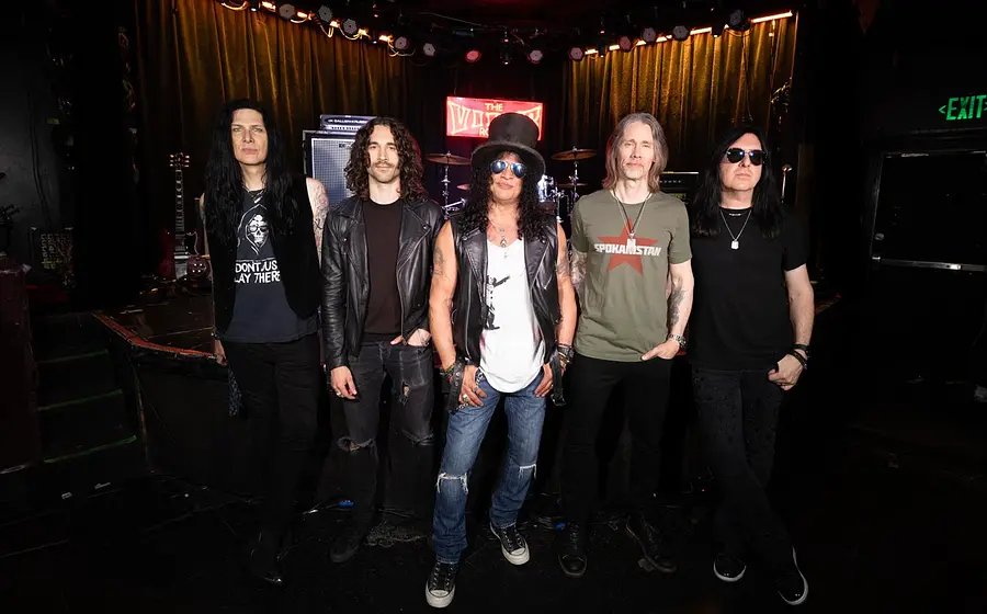 Slash und Myles Kennedy mit drei Personen der Band Conspirators blicken in die Kamera. Im Hintergrund eine Bühne mit Schlagzeug und Gitarre.