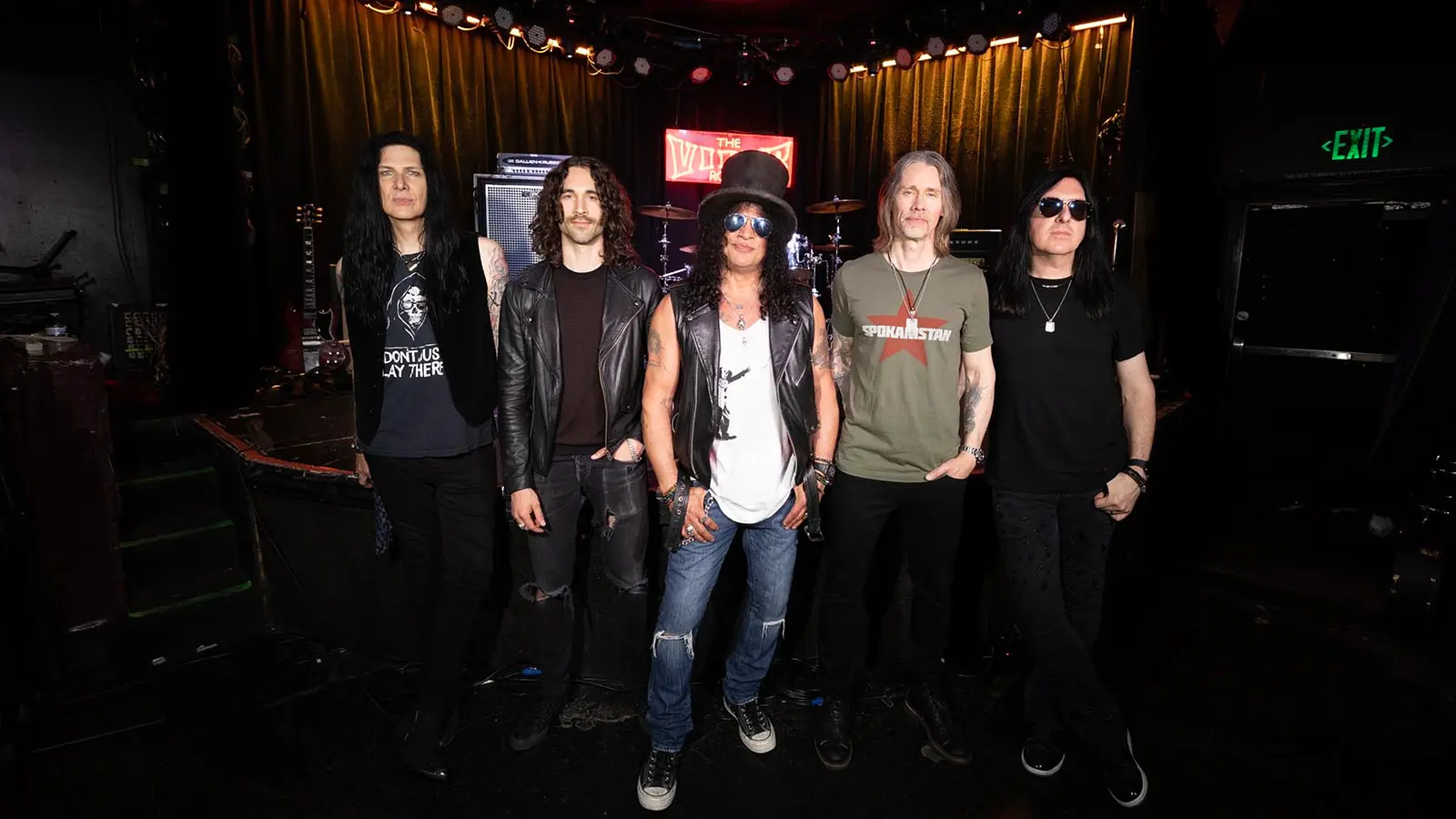 Slash und Myles Kennedy mit drei Personen der Band Conspirators blicken in die Kamera. Im Hintergrund eine Bühne mit Schlagzeug und Gitarre.