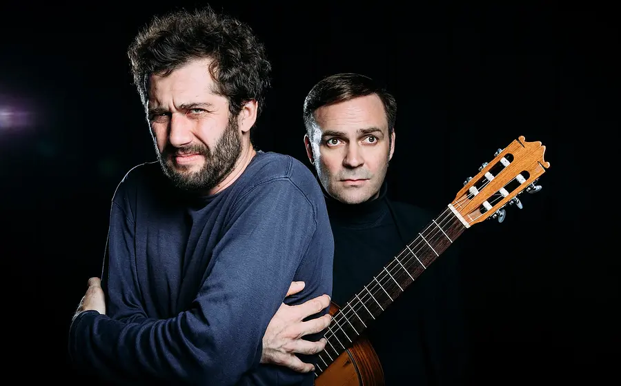 Porträtfoto mit zwei Personen vor schwarzem Hintergrund, eine Person im dunklen Pullover mit verschränkten Armen im Vordergrund, die andere dahinter mit klassischer Gitarre über der Schulter und schwarzem Rollkragenpullover.